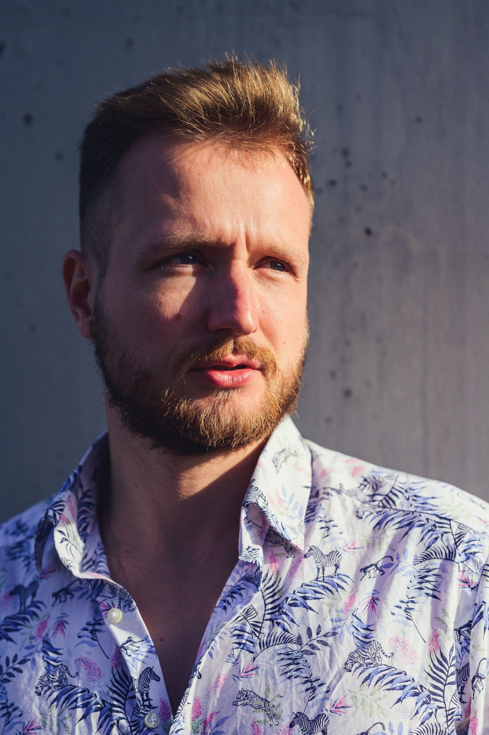 Close-up portrait of a bearded man in a floral shirt against a gray background.